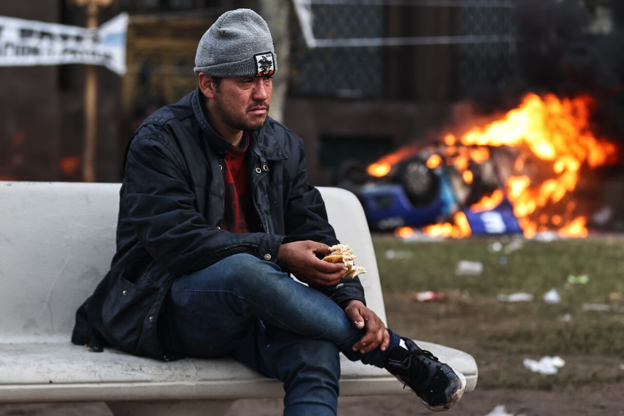 Fotografía de un hombre comiendo con un auto quemado atrás de él durante enfrentamientos entre la policía y personas que protestan a las afueras del senado. Foto: EFE/ Juan Ignacio Roncoroni