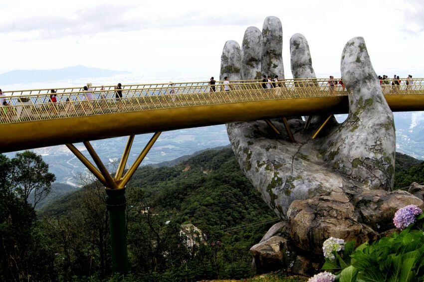 
Foto: AFP | 31 de julio de 2018. Turistas caminan a lo largo del "Puente Dorado" de Cau Vang de 150 metros de largo en las colinas de Ba Na. Acurrucados en las colinas boscosas del centro de Vietnam, dos gigantescas manos de hormigón emergen de los árboles y sostienen un brillante puente dorado.
   