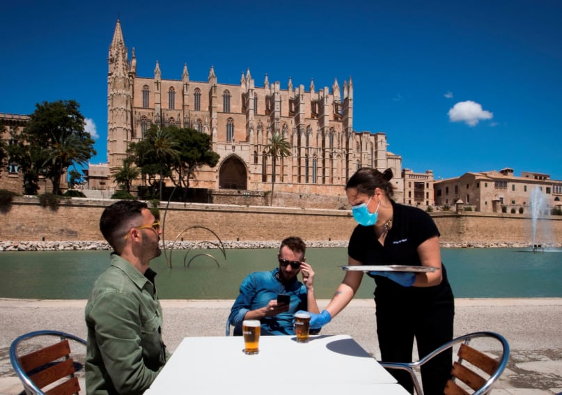 
Una camarera con una máscara facial y guantes sirve a los clientes en un bar con terraza cerca de la Catedral de Palma de Mallorca | AFP
   