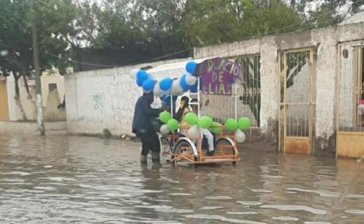 Un padre empuja un carro en medio del agua. Arriba del vehículo va su hijo, quien se recibió y ambos salieron a festejar por las calles.